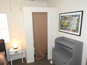 Un dormitorio con cama, mesita de noche con lámpara, cómoda gris, una puerta y un cuadro en la pared.