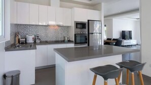 Une cuisine moderne avec des armoires blanches, un comptoir en granit, un four encastré et un réfrigérateur en acier inoxydable. Il y a deux tabourets de bar près de l’îlot central.