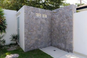 Une cabine de douche en pierre avec un petit seuil à l’entrée, entourée d’un mur blanc et d’une zone herbeuse.
