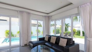 Un salon moderne avec un canapé en cuir noir, une table basse en verre et de grandes fenêtres donnant sur une piscine et un espace verdoyant.