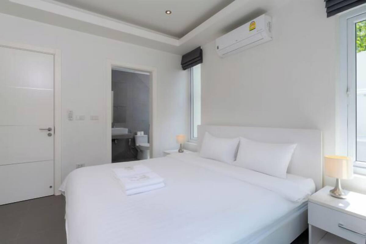 Habitación de hotel con una cama grande, una mesita de noche con lámpara, equipo de aire acondicionado y ventana con vistas a zonas verdes.