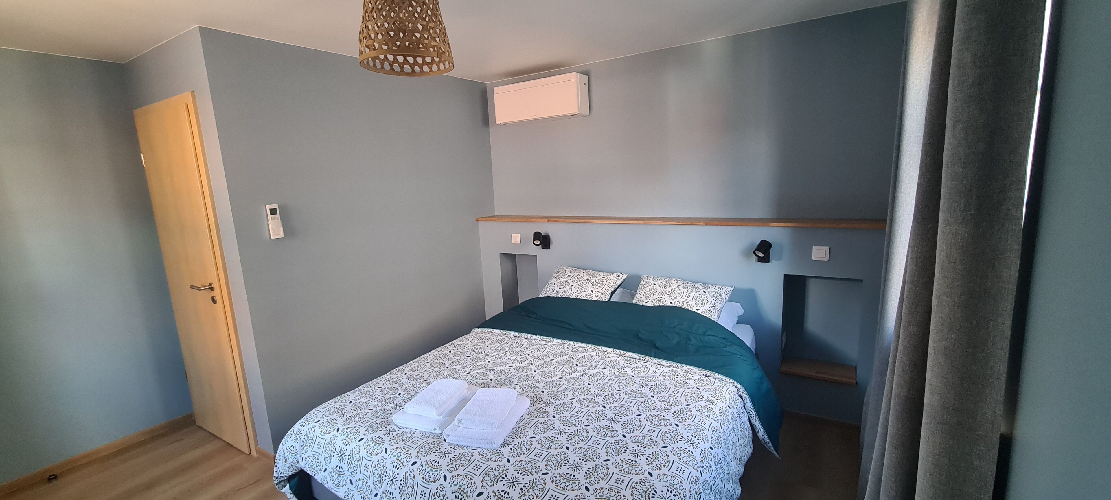 A neatly made bed with a patterned bedspread and pillows, a wooden headboard, a wall-mounted air conditioning unit, and a door on the left.