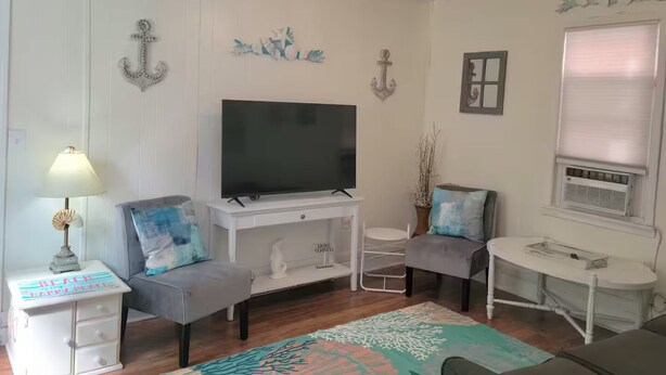 Un salón con televisor de pantalla plana, una mesita, dos sillas, una lámpara, detalles decorativos en la pared y una ventana con persianas.
