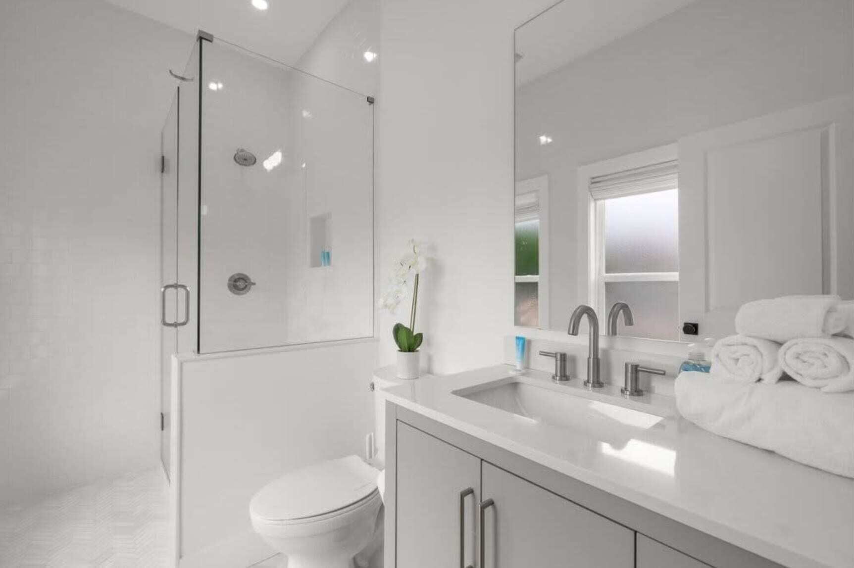 Een moderne badkamer met een glazen douchecabine, toilet in het wit, een wastafel met spiegel en een raam.