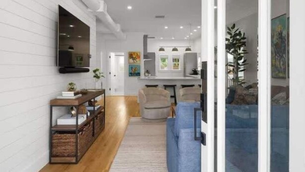 A modern living room with a wooden floor, a blue sofa, a TV mounted on the wall, and a ceiling fan.