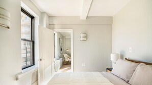 A bedroom with a large bed, a bedside table with lamps, a window, and a door leading to another room. - Upper west side 3 bedroom retreat (New York)