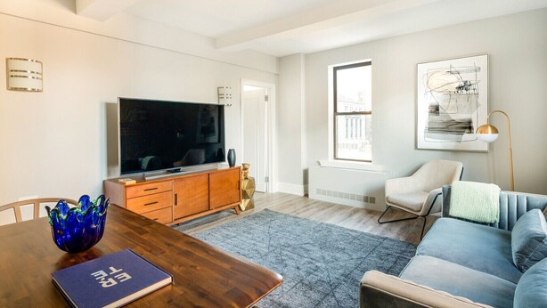 A modern living room with a flat-screen TV, wooden cabinet, blue sofa, and a dining table with a book. - Upper west side 3 bedroom retreat (New York)