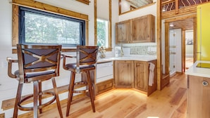 A modern kitchen with wooden cabinets, a central island, and two bar stools. - Tiny in Paradise! Near Chico and SAGE Lodge! (Pray)