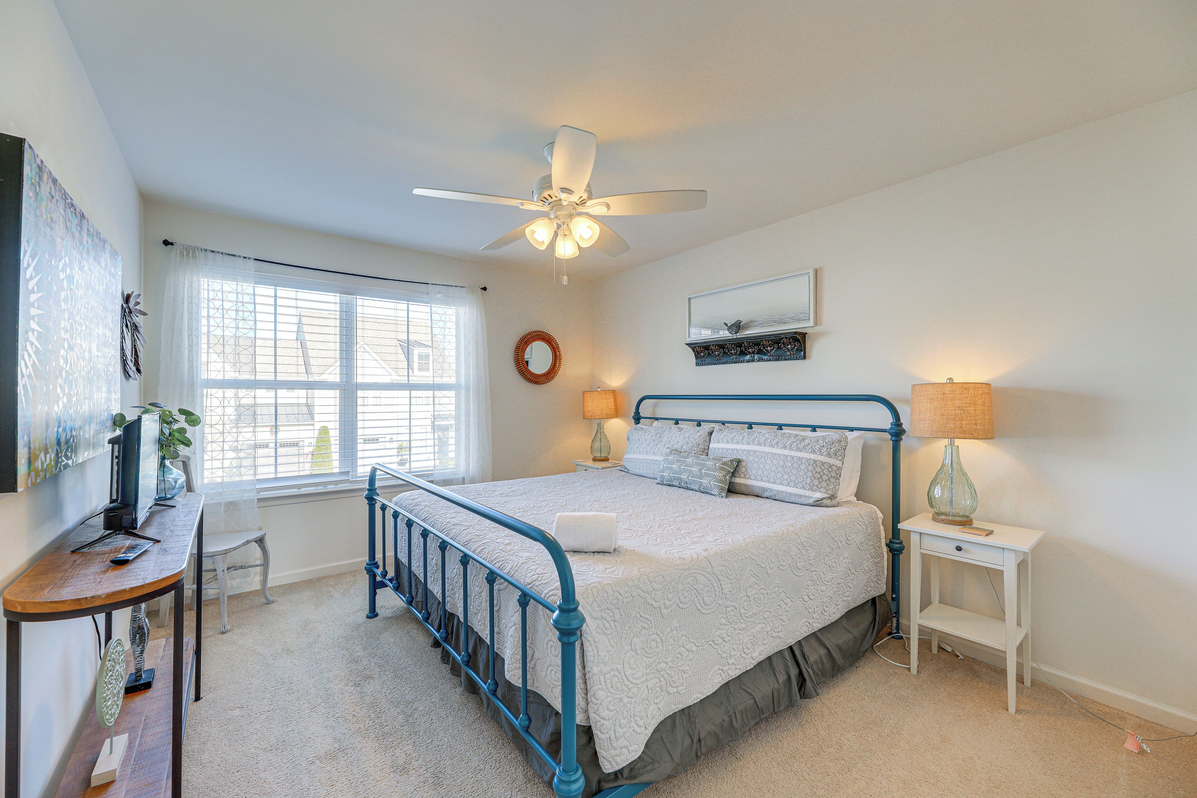 A bedroom with a bed, a desk, a chair, a ceiling fan, a window, and a nightstand with a lamp.