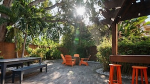 Een terras buiten met een houten tafel, oranje stoelen en een pergola met hangende lampen.