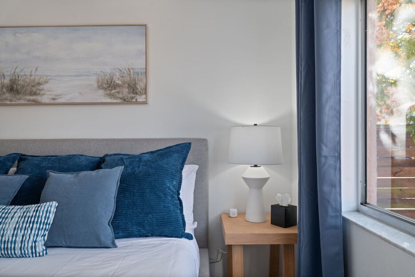 Una cama bien hecha con almohadas azules y blancas, una lámpara de noche y un cuadro con una playa en la pared.
