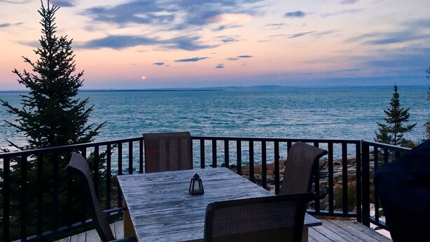 A wooden deck with a table and chairs overlooking a body of water at sunset. - Le P'tit Bijou au Bord du Fleuve (Saint-Siméon)