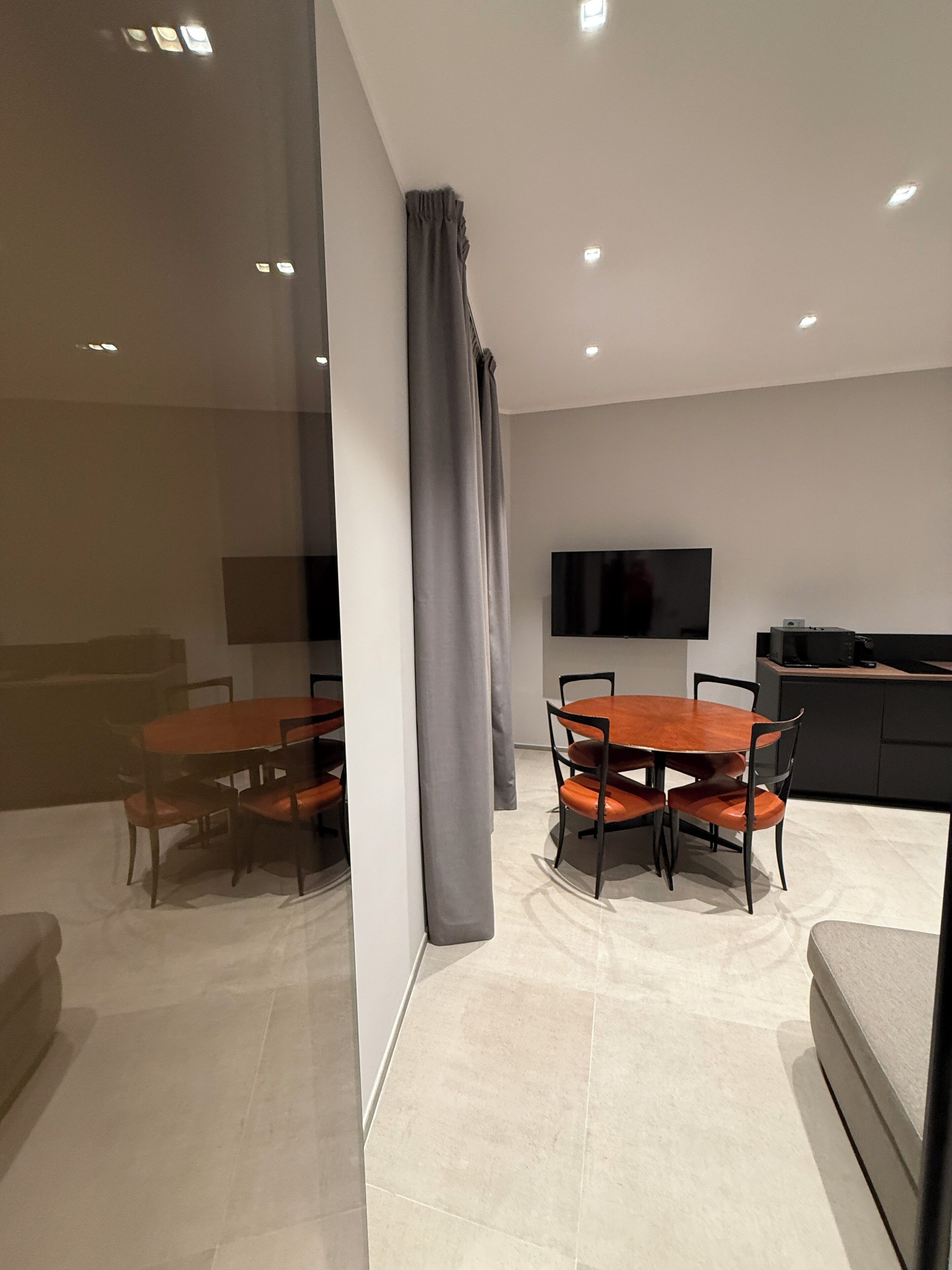 A modern dining area with a round table and chairs, a flat-screen TV mounted on the wall, and a kitchen area with a countertop and appliances.