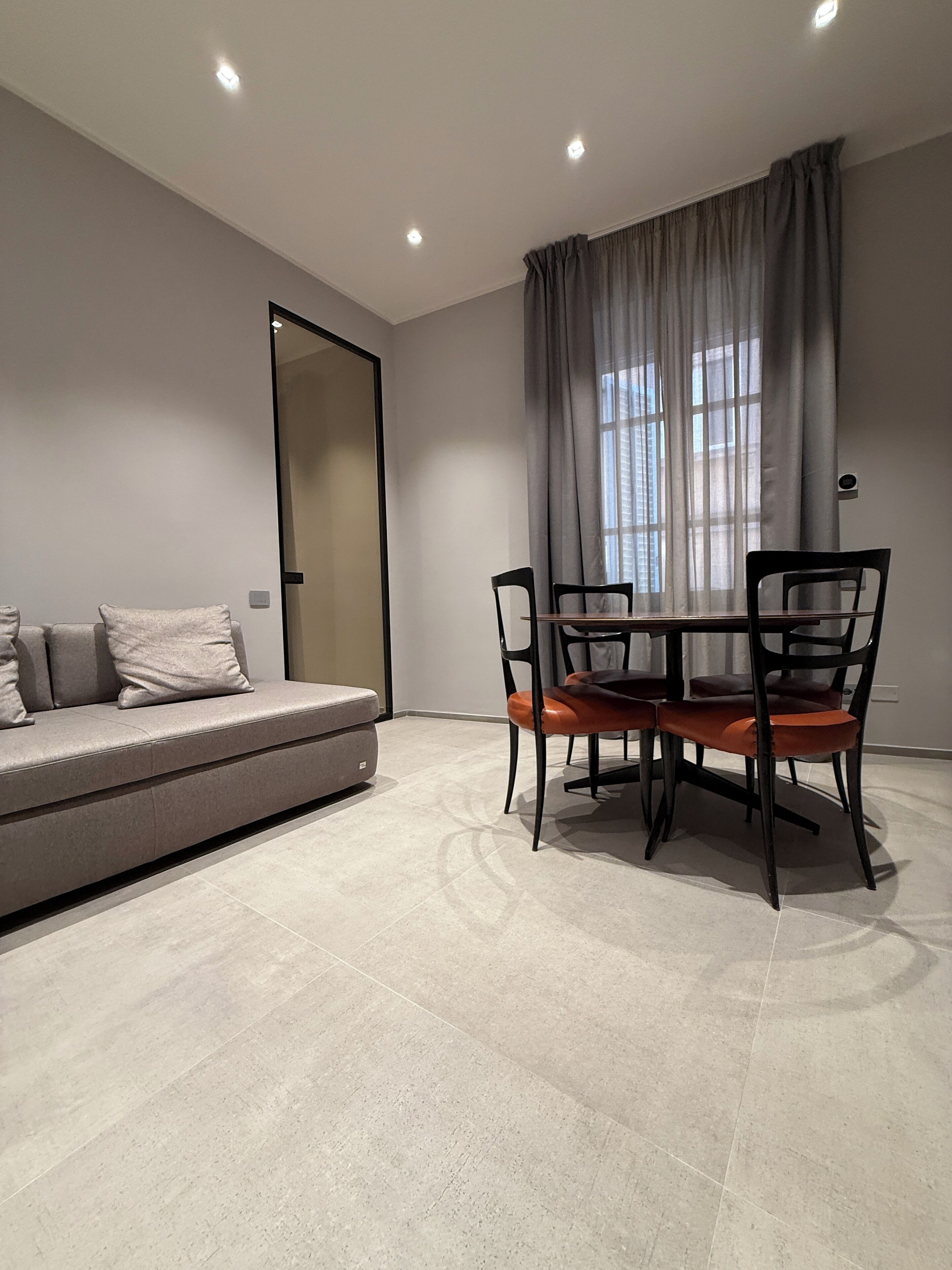 Een moderne woonkamer met een bank, een eettafel met stoelen, een groot raam met gordijnen en inbouwspots.