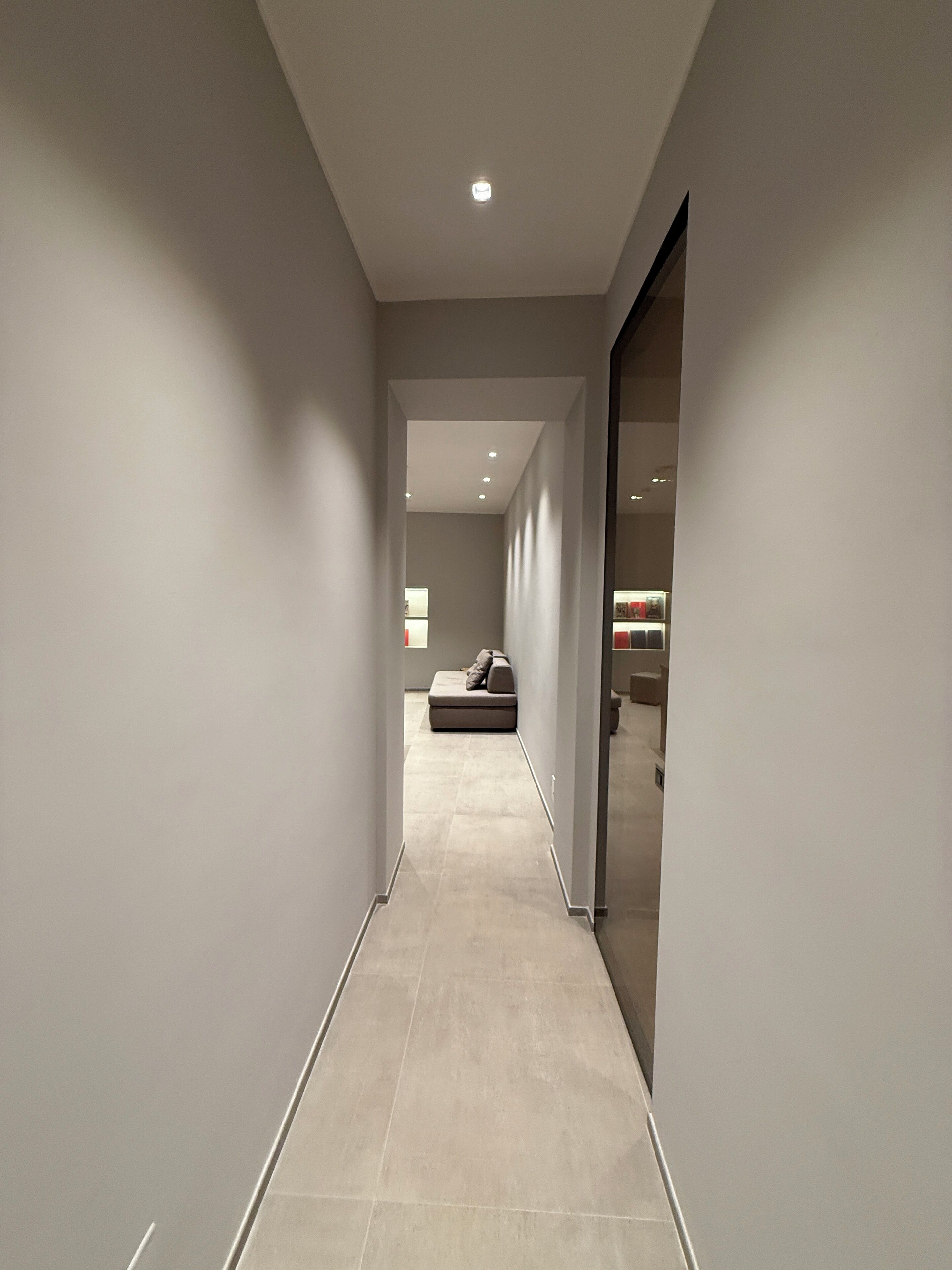 Um corredor estreito e bem iluminado, com cores neutras, que dá acesso a um quarto com cama e uma pequena mesa.