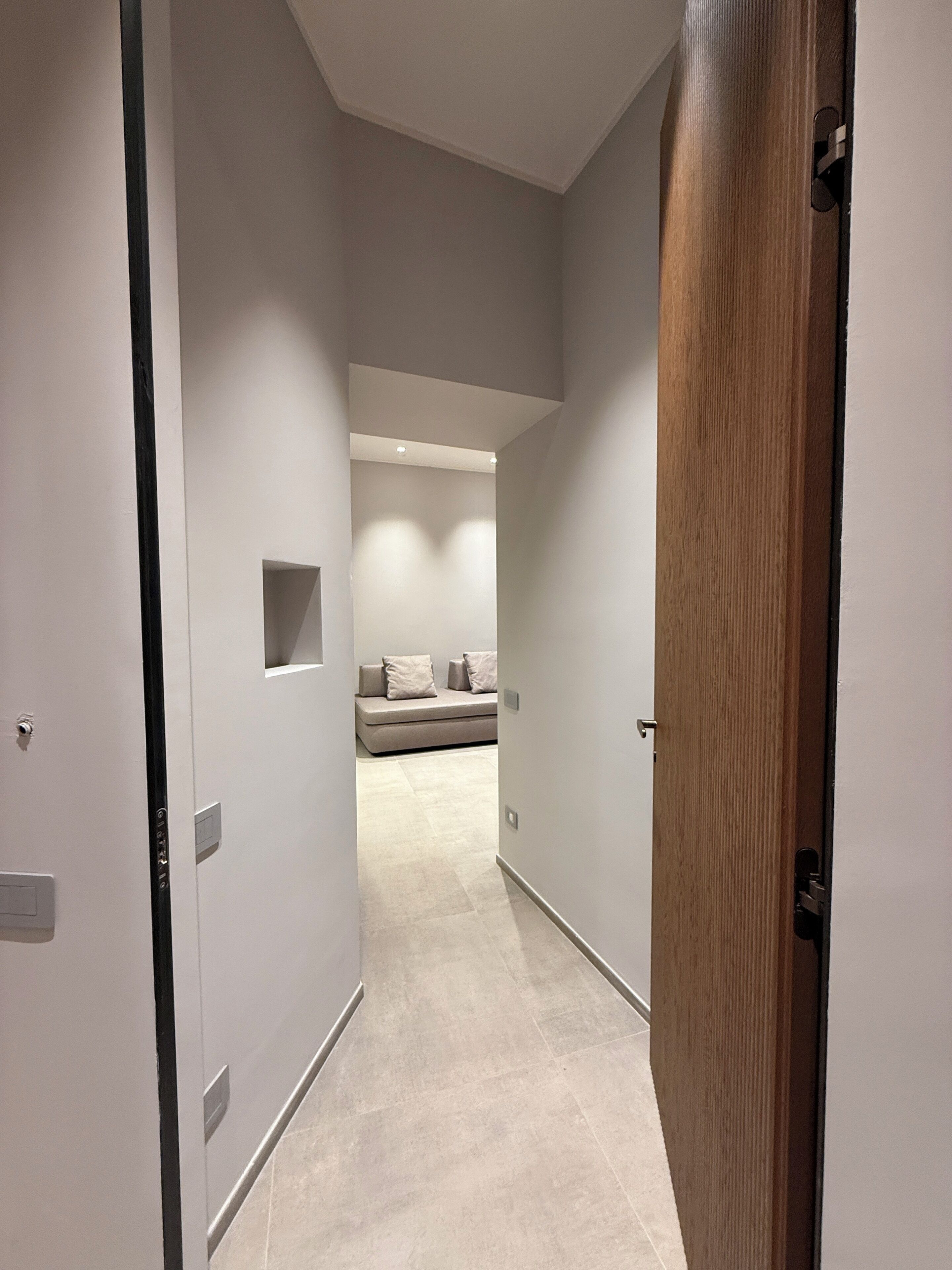 A narrow hallway with a wooden door on the right, leading to a room with a bed and a small window.