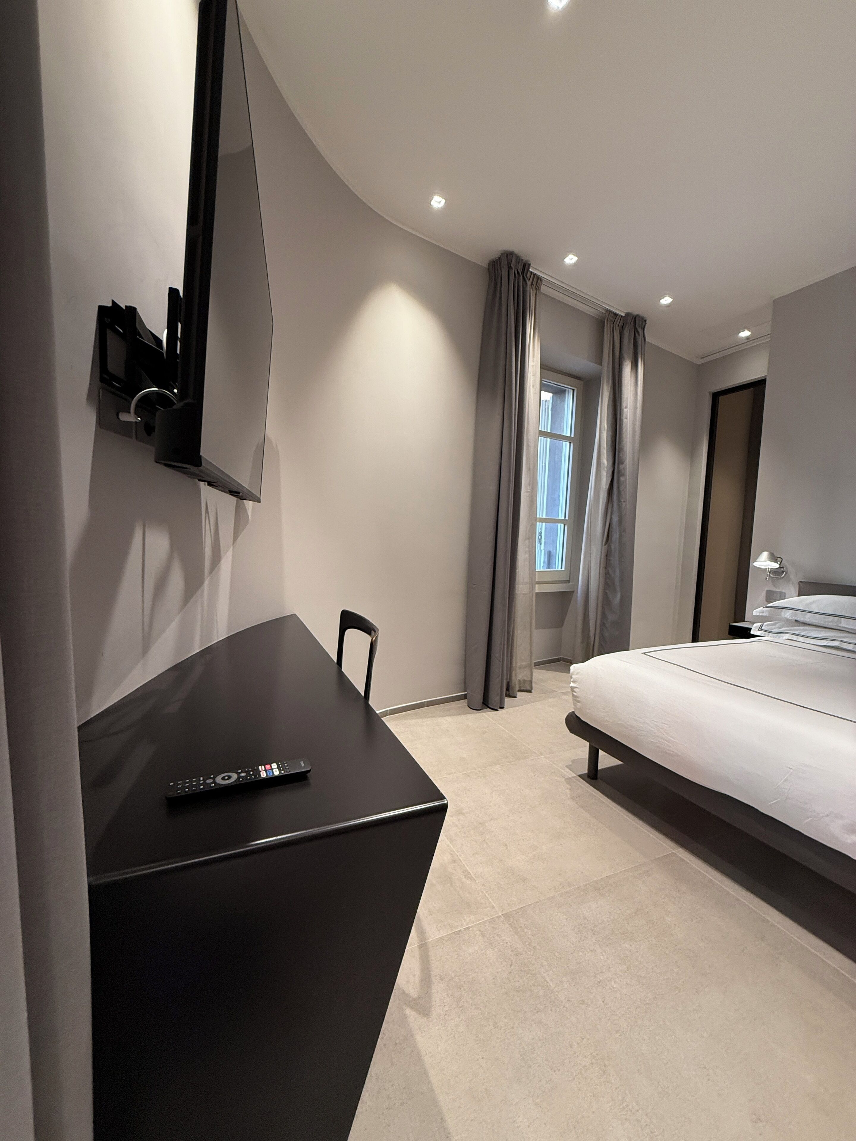 Et moderne hotelværelse med et vægmonteret fladskærms-tv, en seng med hvid madras, et lille skrivebord med en fjernbetjening og et vindue med gardiner.