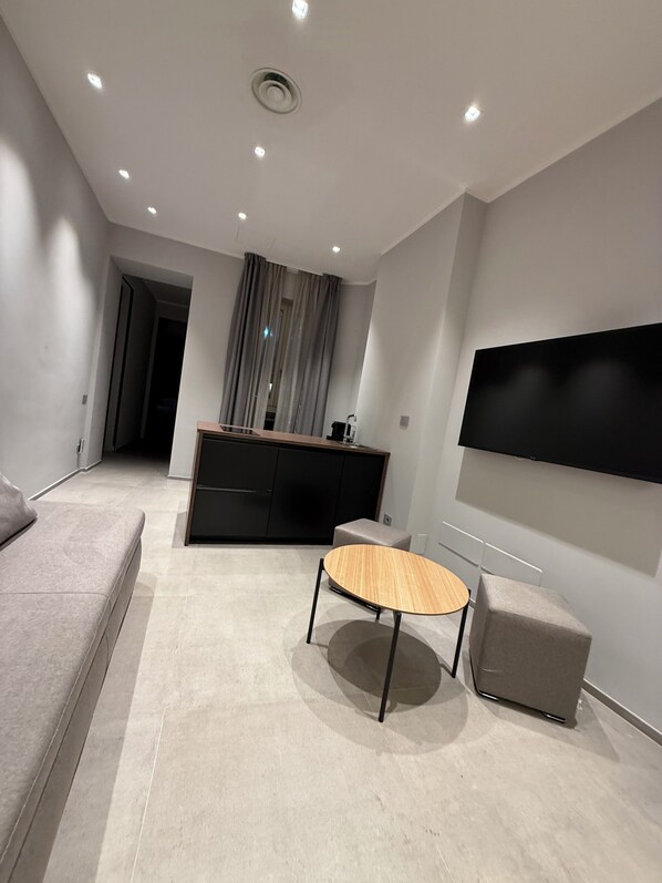 Un salon moderne comprenant un canapé, une table ronde, un téléviseur fixé au mur et un coin cuisine avec un meuble et des rideaux.