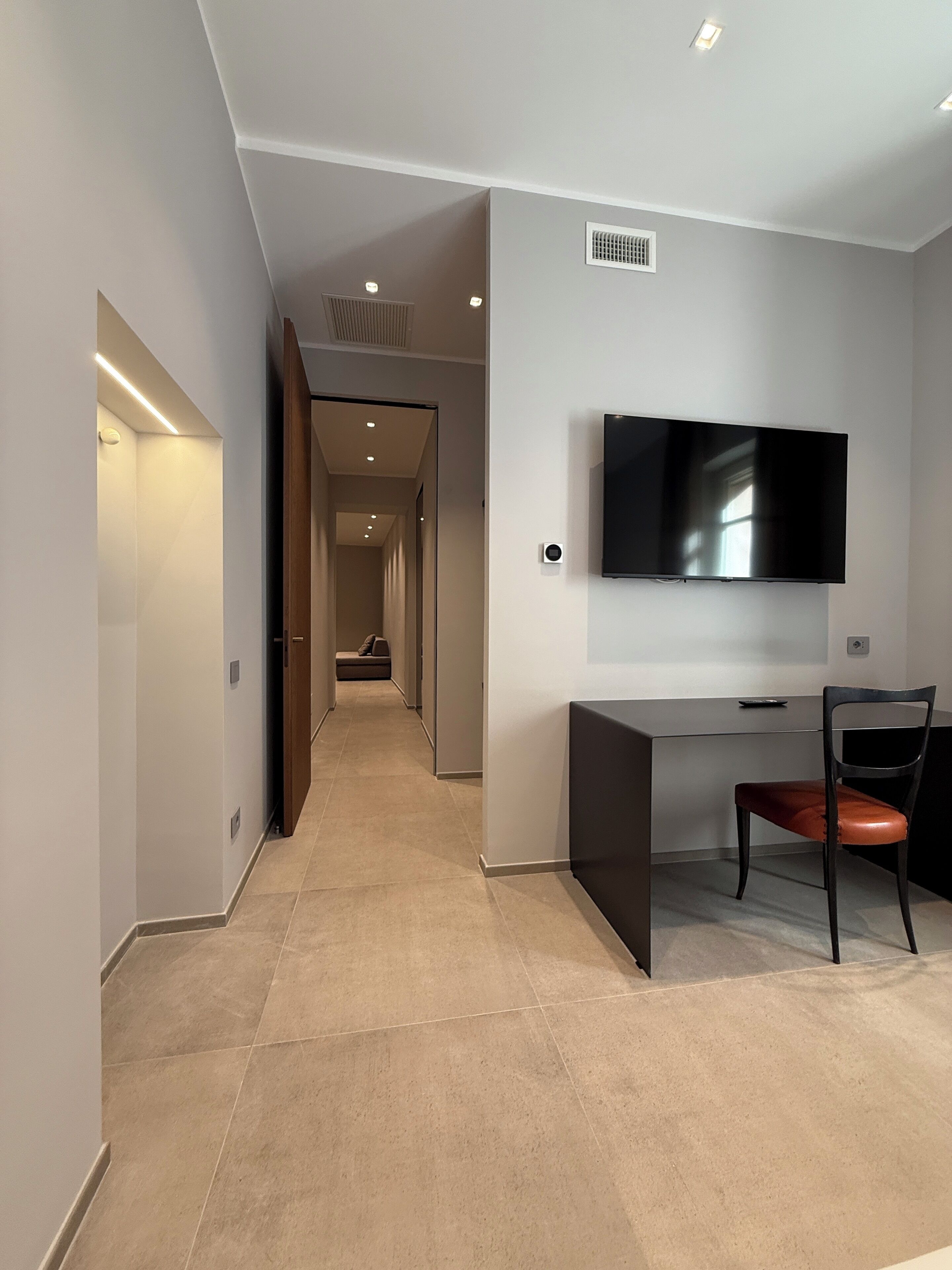 Een moderne hotelkamer met een flatscreen televisie aan de muur, een bureau met een stoel, en een gang naar een andere kamer.