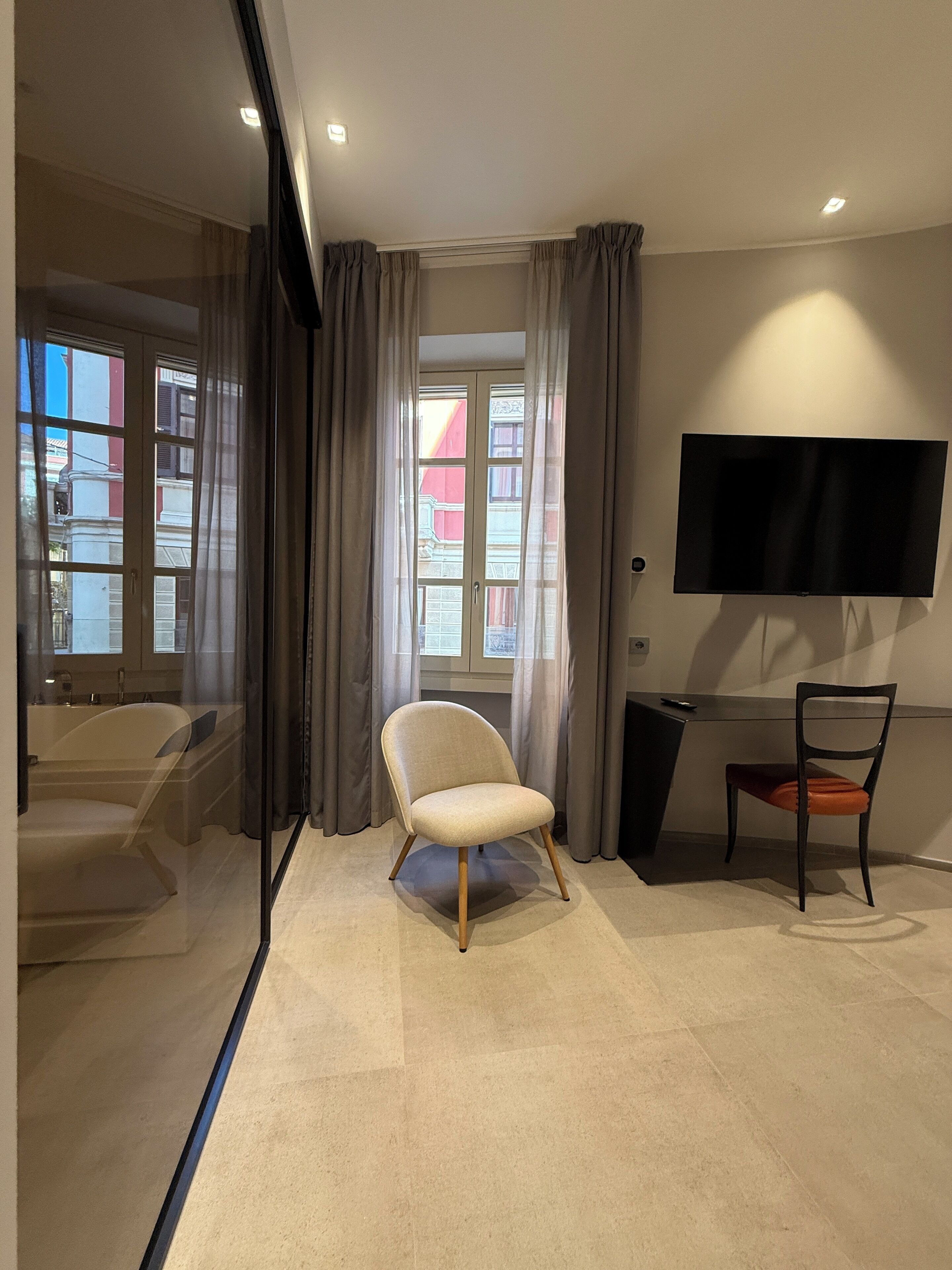 A modern hotel room with a large window, a flat-screen TV mounted on the wall, a chair, and a desk.