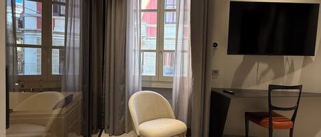 A modern hotel room with a large window, a flat-screen TV mounted on the wall, a chair, and a desk.