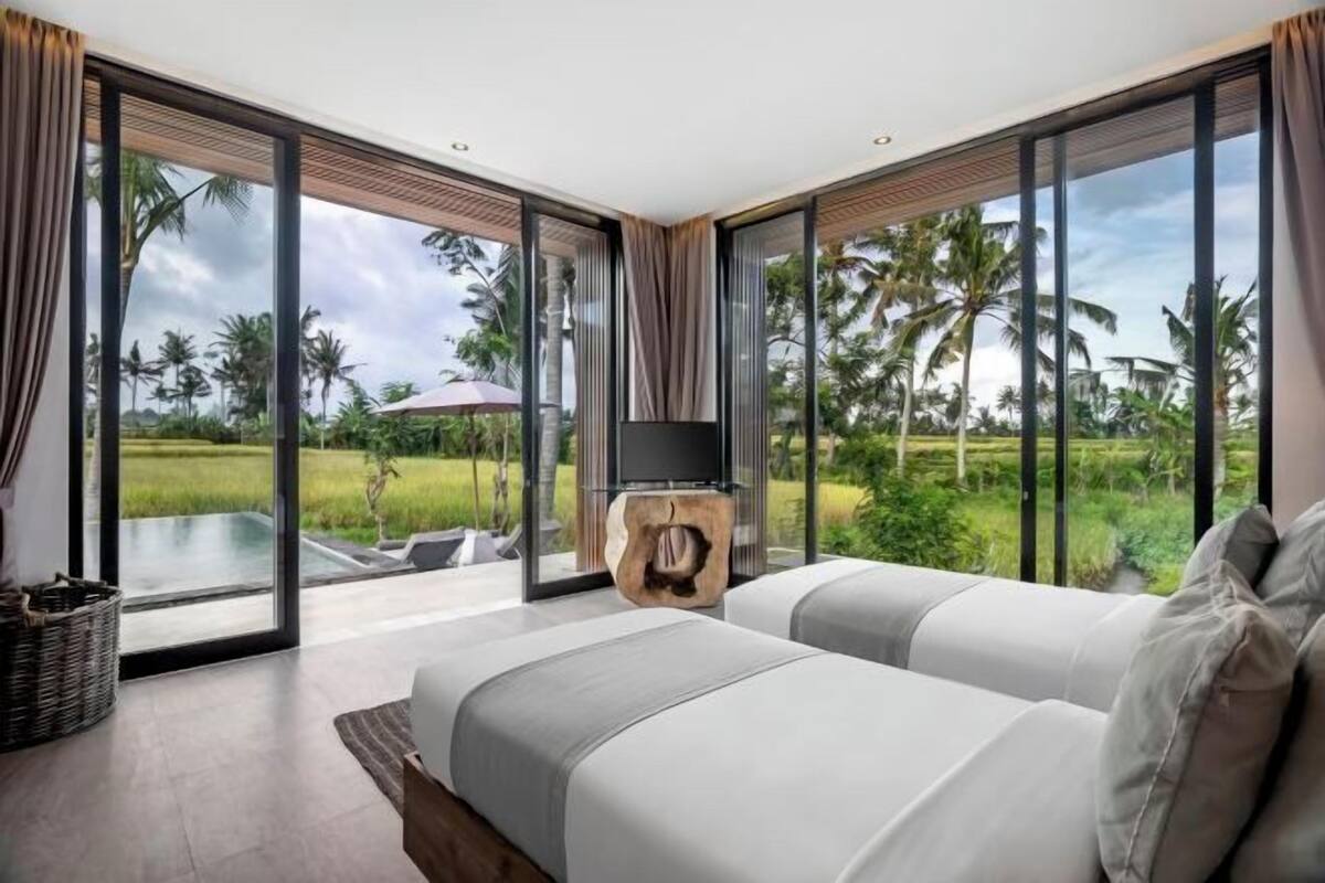 A bedroom with a large window, a bed, a wooden side table, and a view of a pool and palm trees.