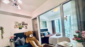 A cozy living room with a large teddy bear, a sofa, a dining table with plates and a vase of flowers, and a sliding glass door leading to a balcony. - Relaxing 1BR Retreat with Balcony (Paranque)