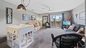 Ein großzügiger Wohnbereich mit einem weißen Tisch, einem beigen Sofa und einem schwarzen Rattan-Sessel. Ein Deckenventilator und eine Pendelleuchte sind zu sehen.