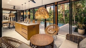 A modern kitchen with a central island, wicker furniture, and a view of an outdoor swimming pool. - Villa Alba by BaliSuperHost (Bali)