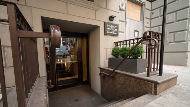 Entrada de hotel con una reja metálica, un cartel que dice "SYNERGY MADISON" y una pequeña maceta en los escalones.