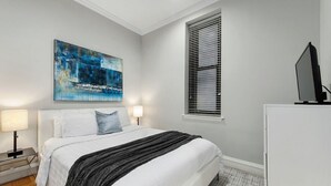 Un dormitorio con cama, mesita de noche, lámpara, televisor, ventana con persianas y un cuadro en la pared.