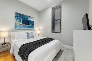A bedroom with a bed, a nightstand, a lamp, a television, a window with blinds, and a painting on the wall.