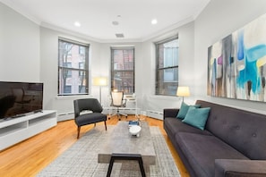 A modern living room with a flat-screen TV, a grey sofa, a black chair, a coffee table, and a desk with a chair.