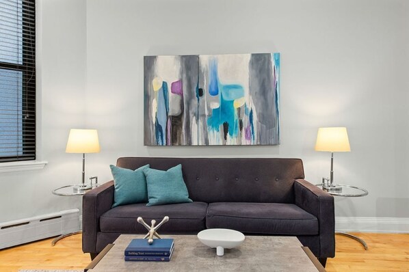 Un salón moderno con un sofá marrón oscuro, cojines color turquesa, una mesa de centro con libros y un objeto decorativo, dos lámparas y un cuadro abstracto grande en la pared.