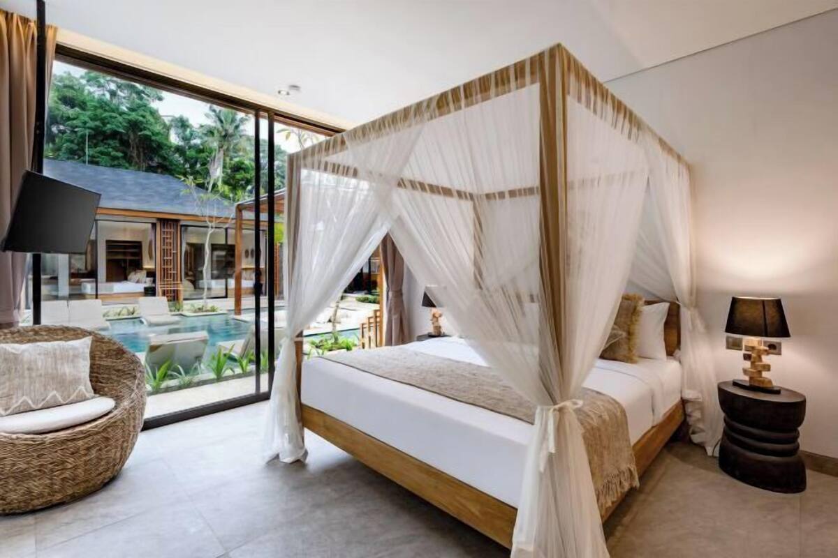 Ein Schlafzimmer mit Himmelbett, Korbsessel, Nachttisch und Blick auf Pool und Außenbereich.