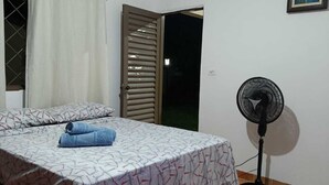 A bedroom with a bed, a standing fan, a window with curtains, and a door. - Namaste Camping Suite (Aruanã)