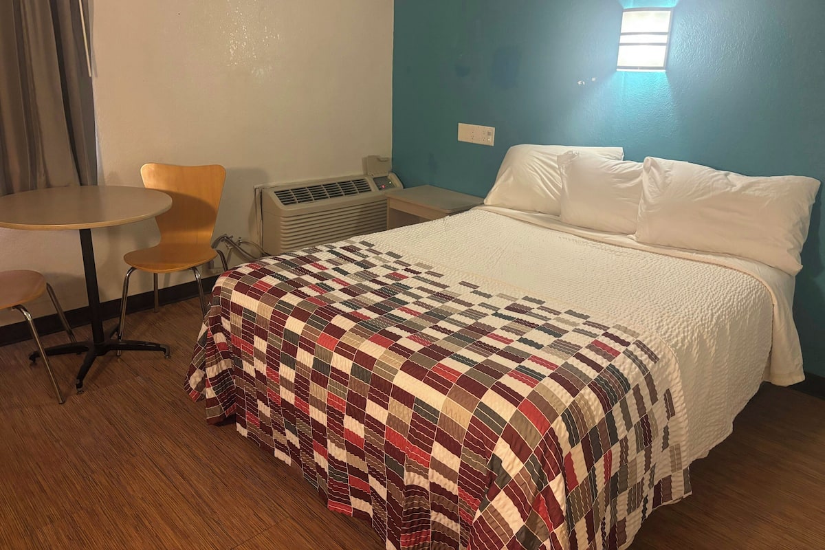 A hotel room with a bed, a chair, a small table, and an air conditioning unit.