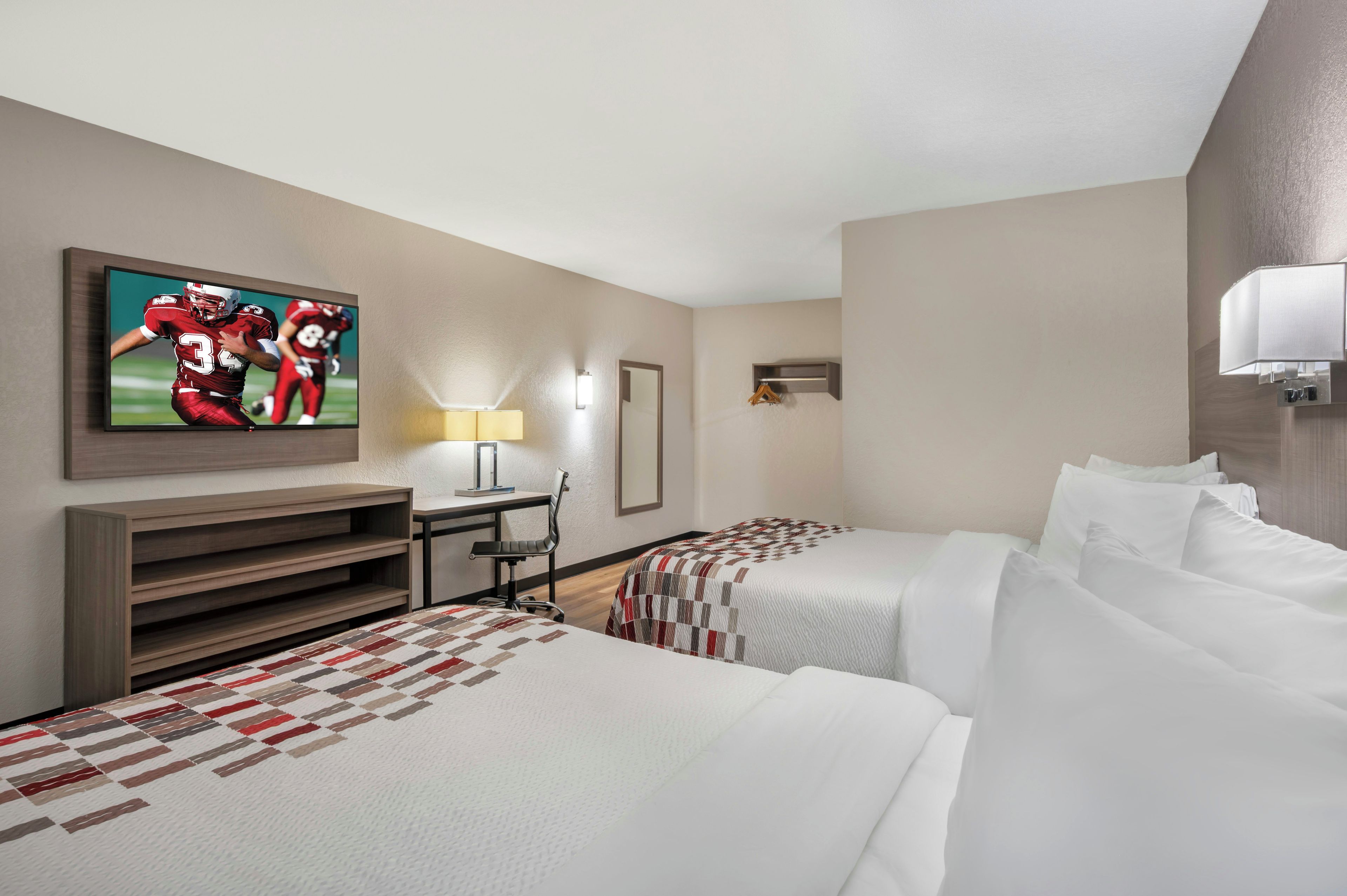 Une chambre d’hôtel avec deux lits, un bureau, une chaise, une télévision et un luminaire fixé au mur.