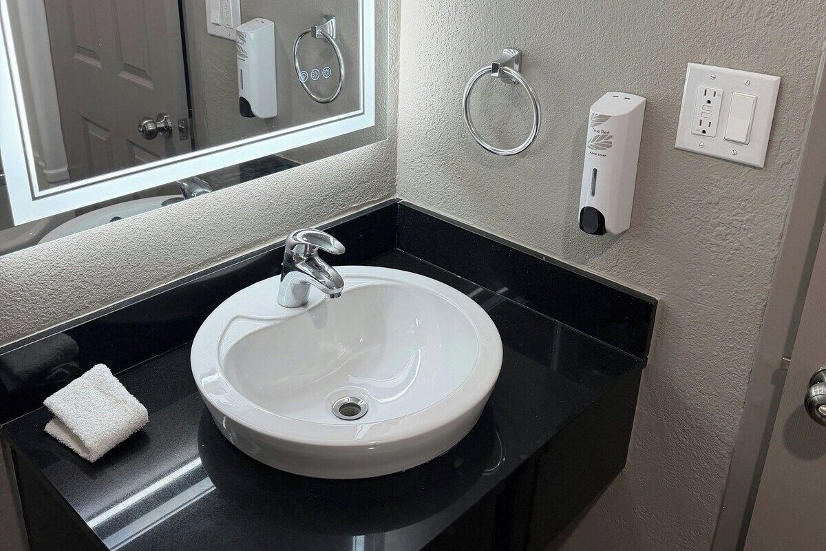 A bathroom with a white sink, black countertop, and a mirror above the sink. There are two towel rings and a soap dispenser on the wall.