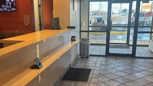 A reception area with a wooden counter, a trash bin, and a view of a parking lot through glass doors. - Red Roof Inn Kokomo (Kokomo)