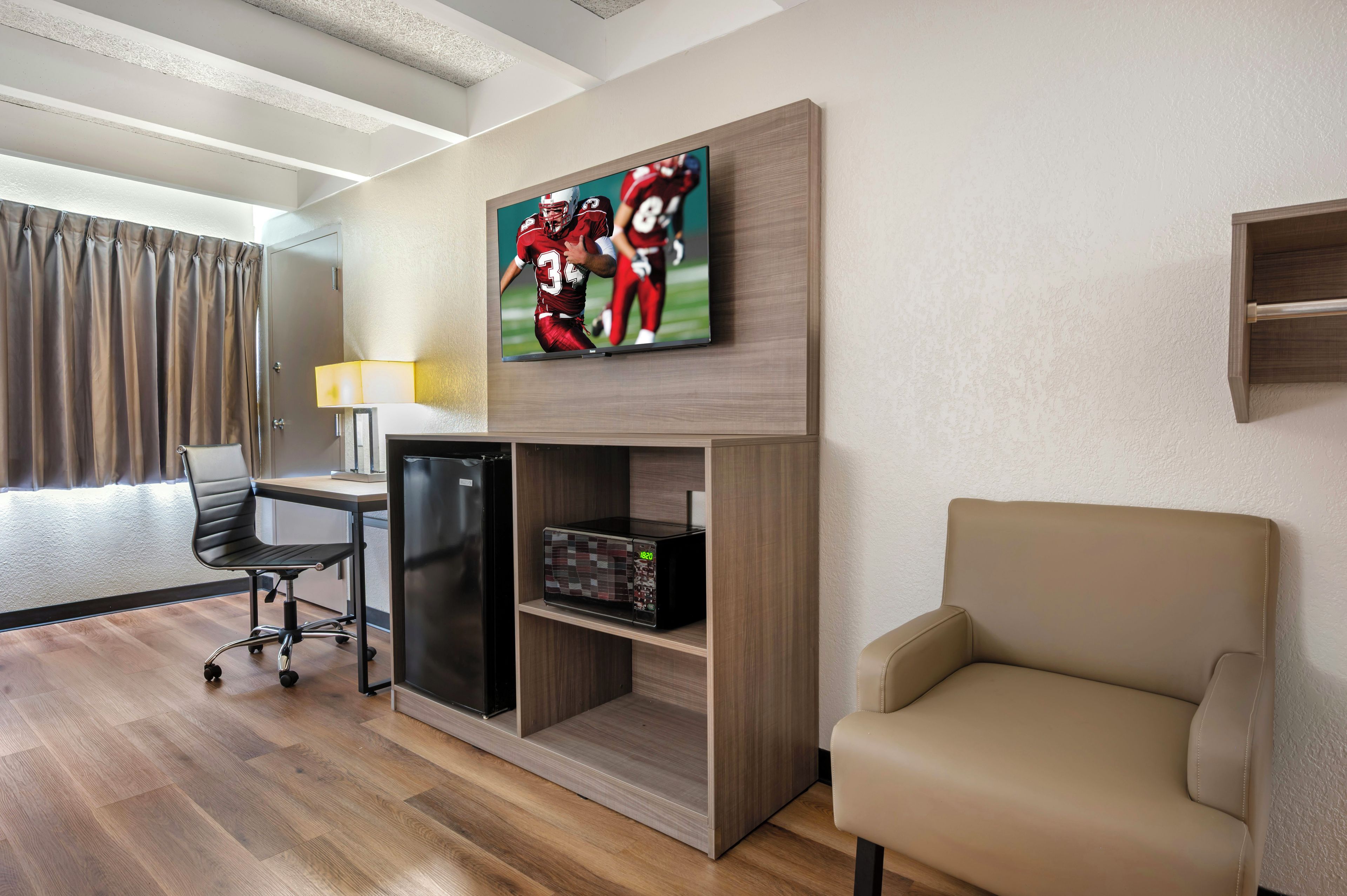 Une chambre d’hôtel équipée d’un téléviseur à écran plat fixé au mur, d’un bureau avec une lampe, d’un réfrigérateur, d’un four à micro-ondes, d’un fauteuil confortable et d’une fenêtre avec des rideaux.