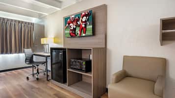 Une chambre d’hôtel équipée d’un téléviseur à écran plat fixé au mur, d’un bureau avec une lampe, d’un réfrigérateur, d’un four à micro-ondes, d’un fauteuil confortable et d’une fenêtre avec des rideaux.