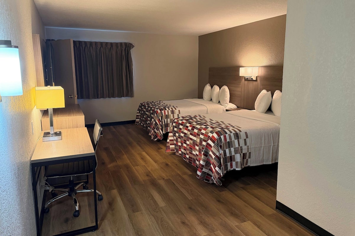 A hotel room with two beds, a desk with a chair, a lamp, and a window with curtains.
