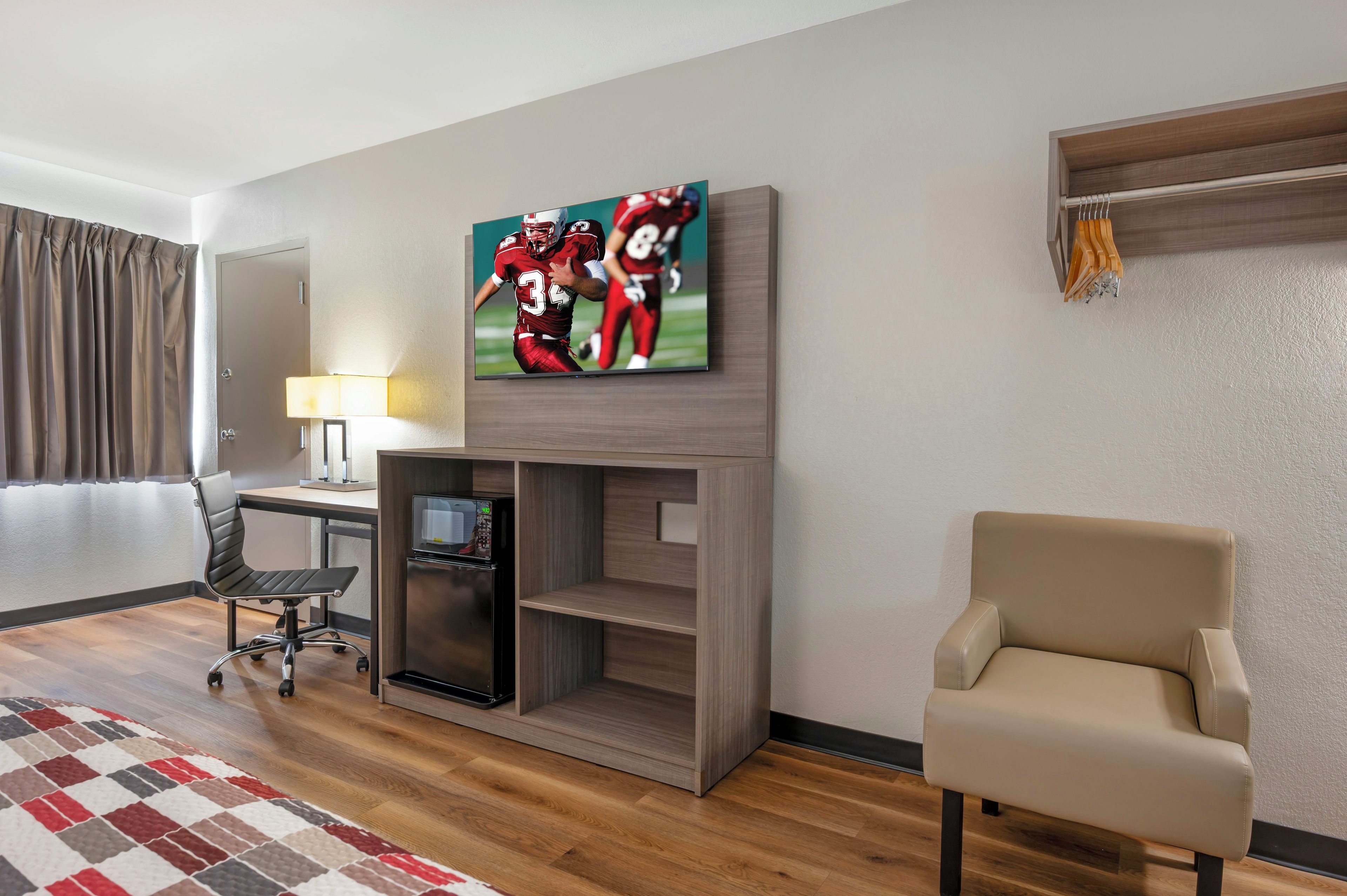 A hotel room with a TV mounted on the wall, a desk with a lamp, a chair, and a wooden floor.