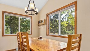 A dining area with a wooden table and chairs, large windows with a view of trees, and a chandelier.