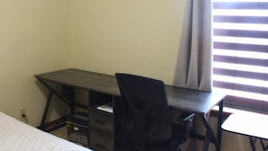 Ein kleines Zimmer mit einem Einzelbett, einem Schreibtisch und einem Stuhl, einem Fenster mit Jalousien und einer Steckdose.