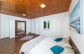 Ein Schlafzimmer mit Holzbalkendecke, einem großen Bett mit gemusterten Kissen, einem Fenster mit Vorhängen und einem Bild an der Wand.