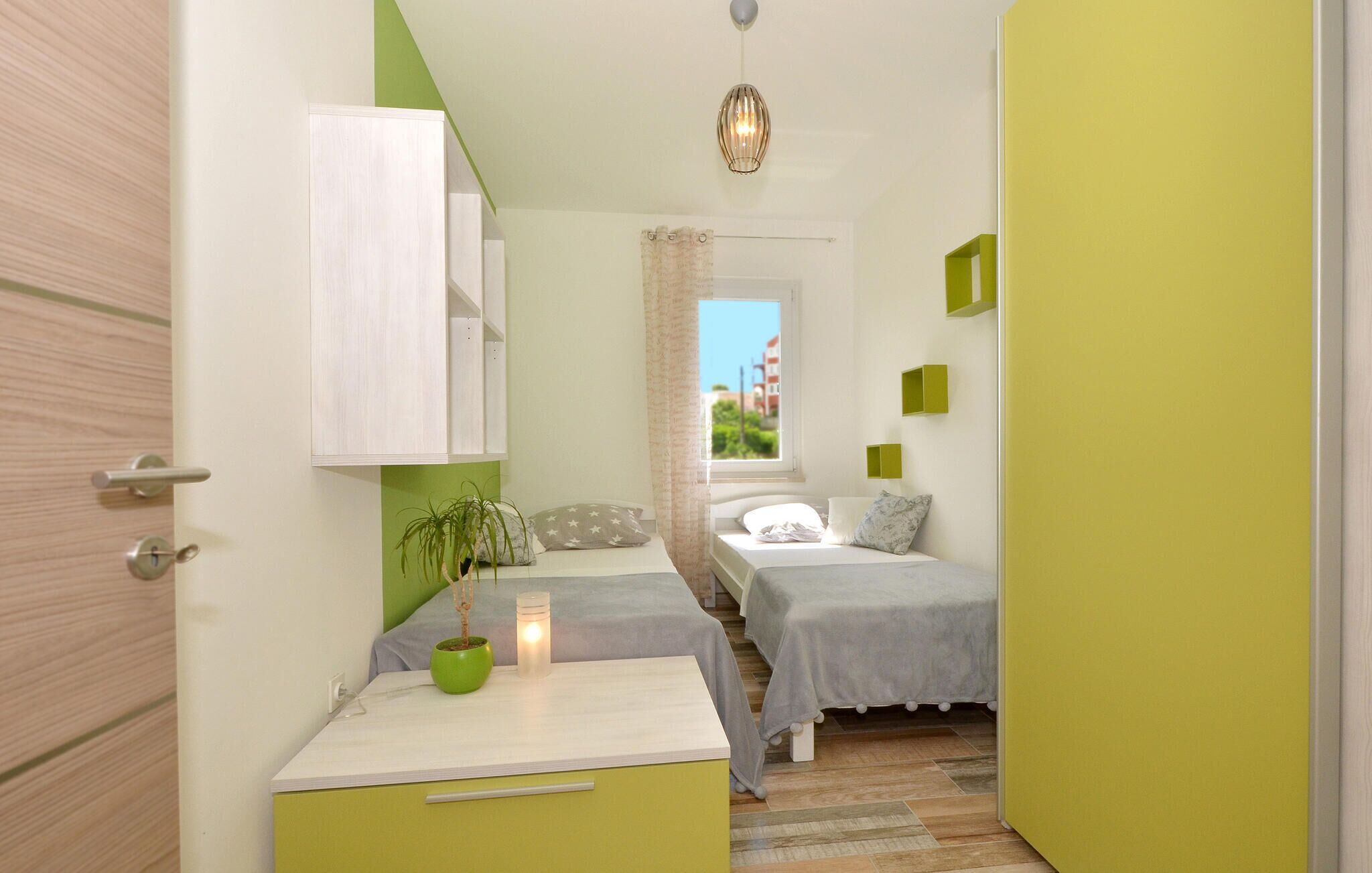 Un dormitorio pequeño y de colores vivos con dos camas, una mesita de noche, una planta en maceta y una ventana que da a vistas de edificios.