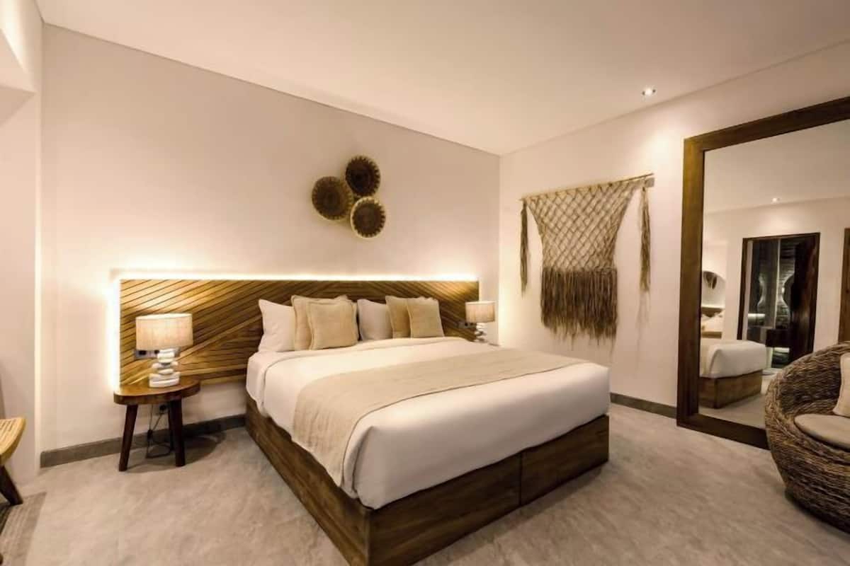 A modern hotel room with a large bed, a bedside table with a lamp, a wall-mounted mirror, and decorative wall art.