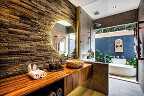 A modern bathroom with a stone wall, wooden vanity, and a large mirror. - Villa Jungle Rendez Vous by BaliSuperHost (Bali)